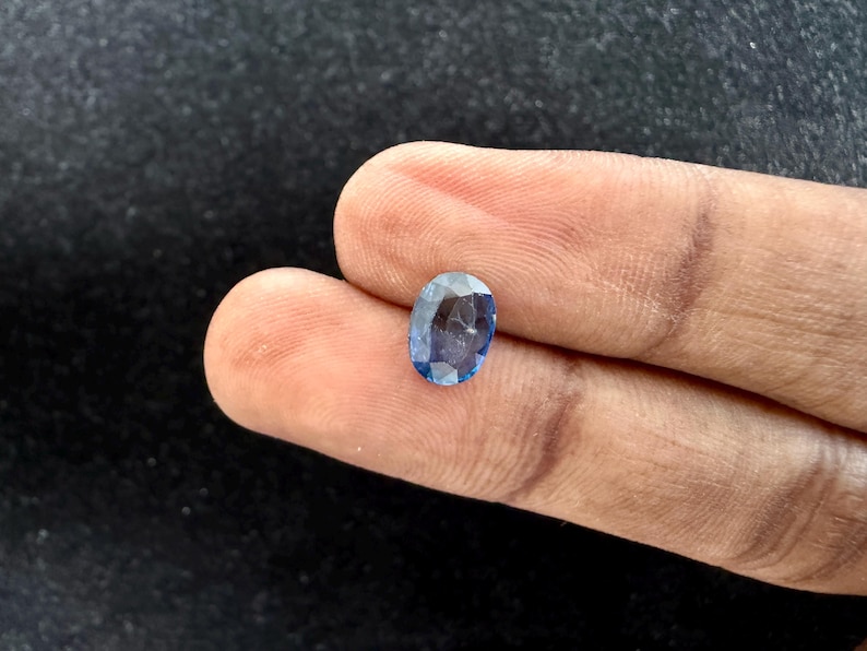 May include: An oval-cut gemstone with a faceted surface and a translucent blue color. The stone displays varying shades of light and dark blue throughout its structure, reflecting light across its polished, angular surfaces.