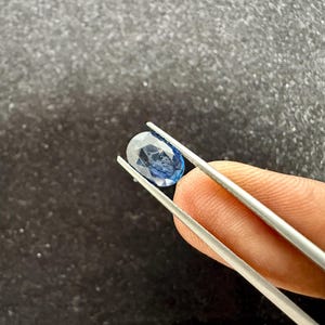 May include: A faceted oval-shaped blue sapphire gemstone held by silver-toned metal tweezers. The gemstone displays a translucent deep blue color with visible internal inclusions and a polished surface reflecting light from multiple angles.