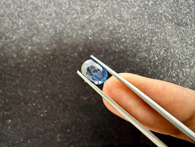 May include: A faceted oval-shaped blue sapphire gemstone held by silver-toned metal tweezers. The gemstone displays a translucent deep blue color with visible internal inclusions and a polished surface reflecting light from multiple angles.