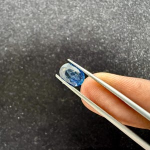 May include: A faceted oval-shaped blue sapphire gemstone held by a pair of silver-toned metal tweezers. The gemstone displays a deep blue hue with visible internal inclusions and a polished surface, reflecting light across its multiple cut facets.