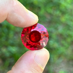 May include: A deep red, faceted gemstone with a starburst pattern. The gemstone is held in a hand, with a green background.