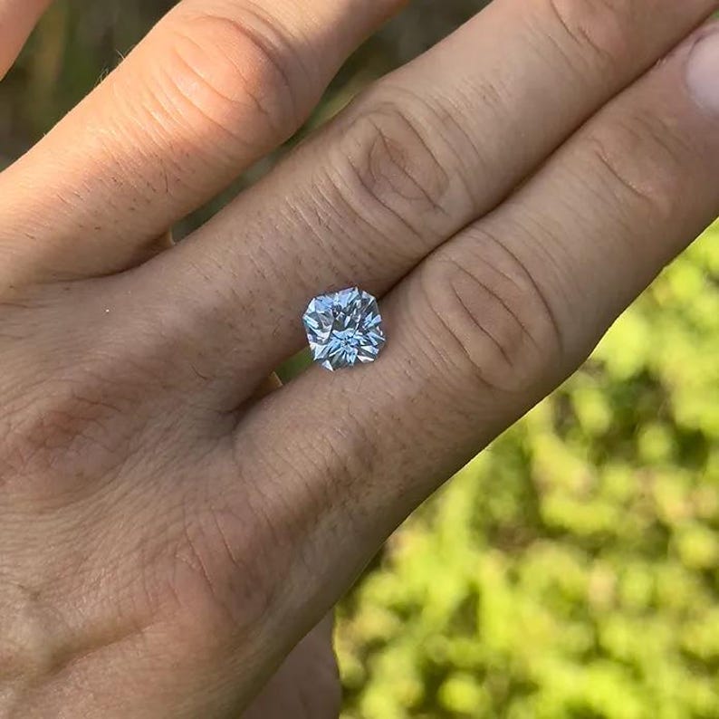 May include: A clear, faceted, square-shaped gemstone with a slight blue hue. The gemstone is set on a finger.