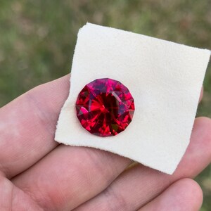 May include: A deep red gemstone with a faceted cut, set on a white fabric background.