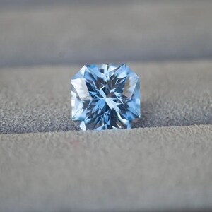 May include: A light blue gemstone with a square shape and many facets. The gemstone is sitting on a gray surface.