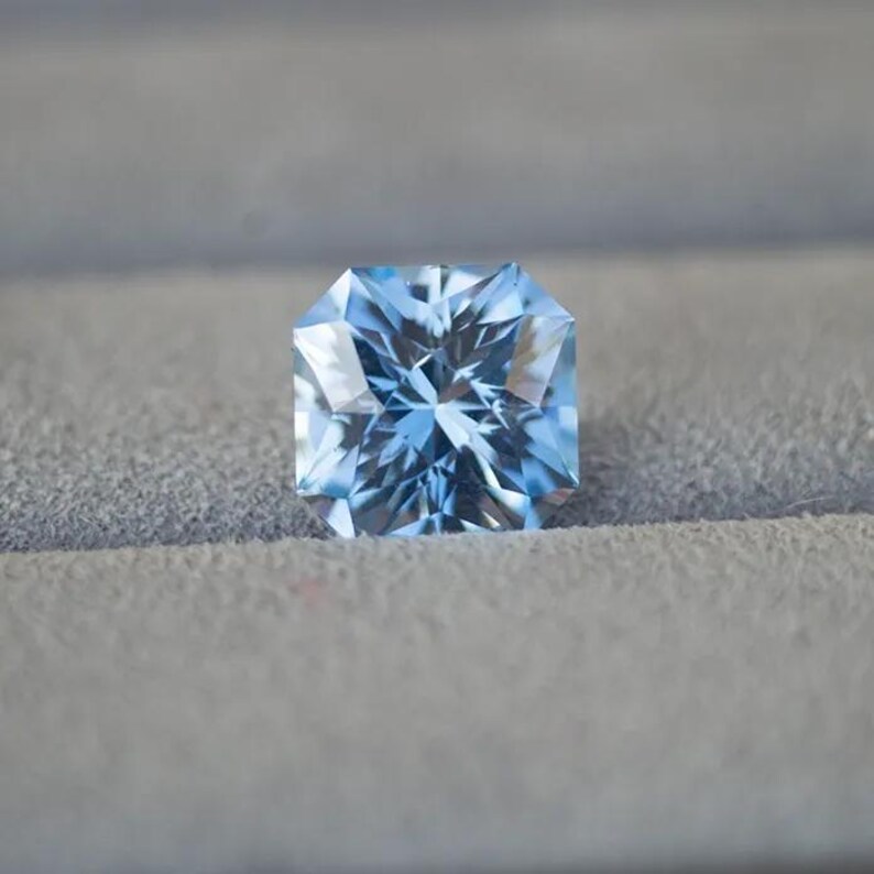 May include: A light blue gemstone with a square shape and many facets. The gemstone is sitting on a gray surface.