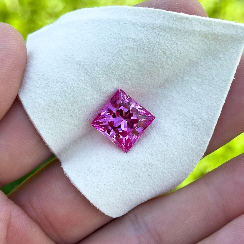 May include: A square-cut, vivid pink gemstone, possibly a sapphire, is displayed on a white cloth. The gem's facets catch the light, highlighting its sparkle. The background is blurred, implying an outdoor location.