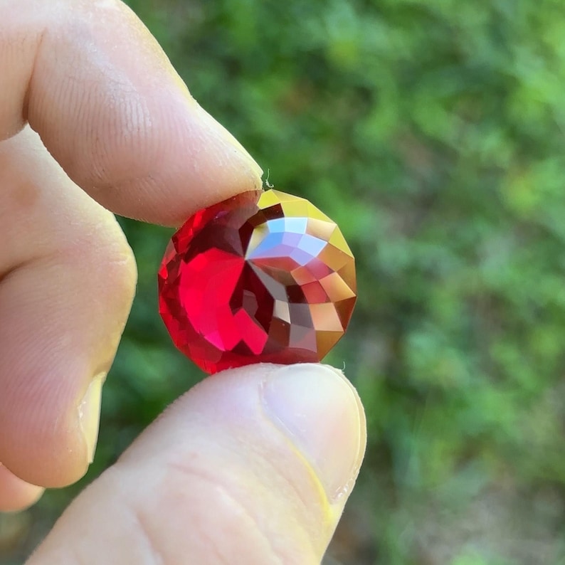 May include: A deep red faceted gemstone with a high shine. The gemstone is held in a hand.