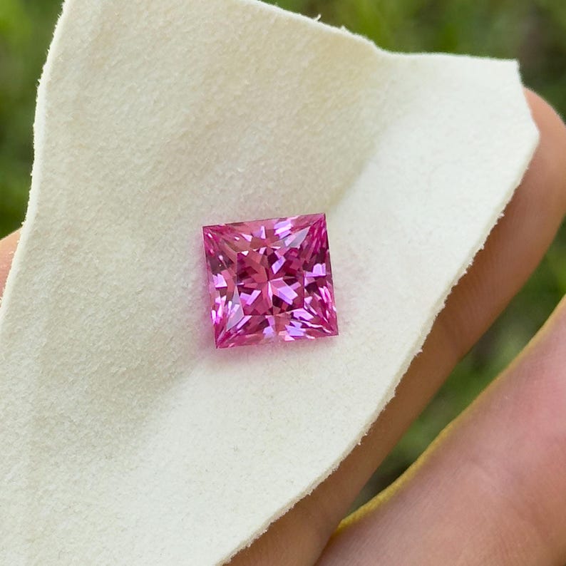 May include: A square-cut, vibrant pink gemstone with multiple facets, resting on a white cloth. The gemstone's colour ranges from light to deep pink, reflecting light. The stone is held by a person.