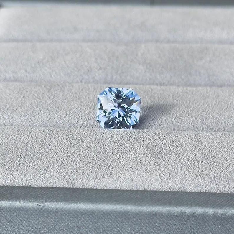 May include: A light blue square-shaped gemstone with facets, set on a gray velvet background.