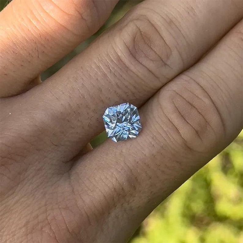 May include: A clear, square-shaped gemstone with a cushion cut. The gemstone is set in a ring and is being worn on a finger.