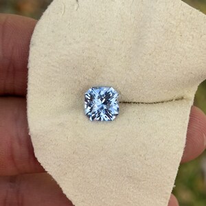 May include: A light blue square-shaped gemstone with facets, displayed on a white fabric surface.