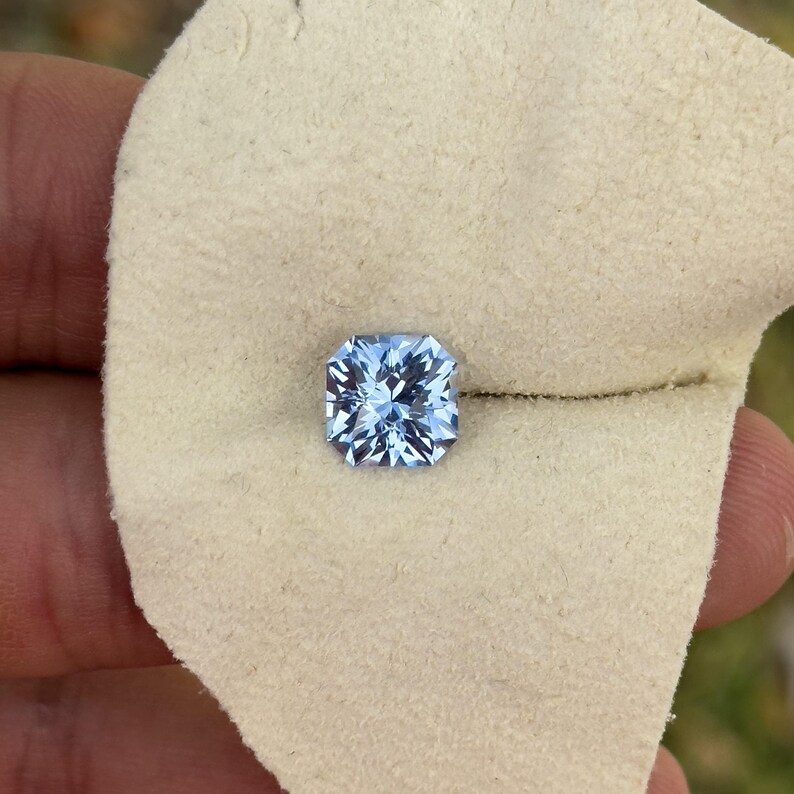 May include: A light blue square-shaped gemstone with facets, displayed on a white fabric surface.
