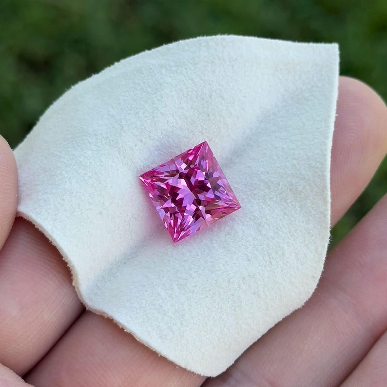 May include: A square-cut, bright pink gemstone is displayed on a white cloth. The gemstone's facets catch the light, highlighting its sparkle. The cloth offers a soft backdrop to the gem's defined edges. The image suggests a focus on jewellery or precious stones.