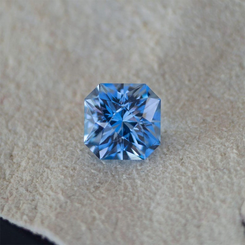 May include: A square-shaped, light blue gemstone with a faceted cut. The gemstone is set against a light brown background.