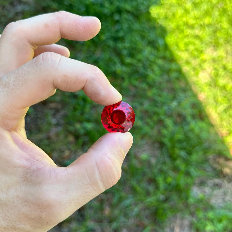 May include: A deep red, faceted crystal gemstone with a round shape.