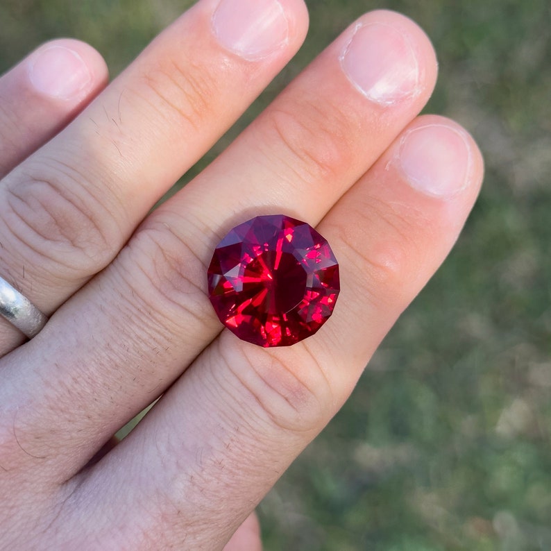 May include: A deep red gemstone with a faceted cut, set in a ring on a person's finger.