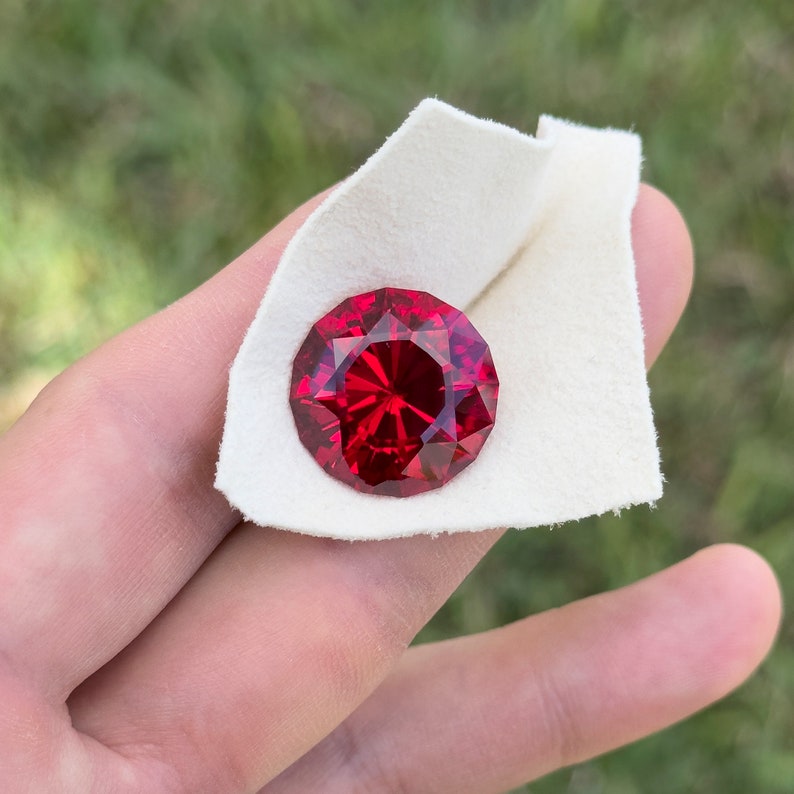 May include: A deep red gemstone with a faceted cut, displayed on a white fabric background.