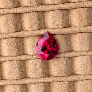 May include: A deep red pear-shaped gemstone on a brown suede background.