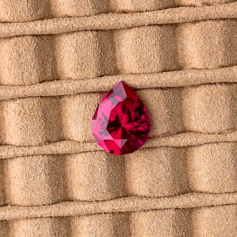 May include: A deep red pear-shaped gemstone on a brown suede background.
