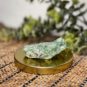 May include: A rough, green stone with flecks of red rests on a small, round, gold-colored stand. The stone has a textured surface and is set against a blurred background of green foliage. The stand sits on a woven, brown surface.