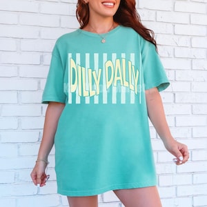 May include: A turquoise t-shirt with the text "Dilly Dally" in a yellow, stylized font. The text is set against a background of vertical white stripes. The shirt has short sleeves and a relaxed fit.
