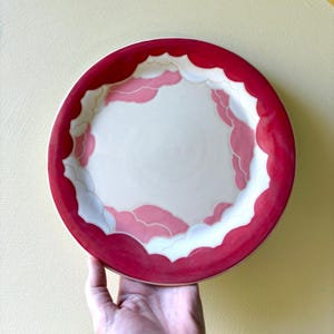 May include: A round ceramic plate with a cream-colored center and a vibrant red rim. The plate features a cloud-like design in shades of pink and white, adding a whimsical touch to the tableware.