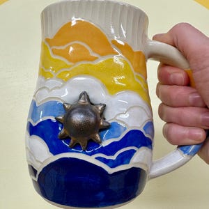 May include: A ceramic mug with a hand-painted design of a sunset over the ocean. The mug features a textured surface, a handle, and a bronze sun-shaped embellishment. The color palette includes shades of orange, yellow, blue, and white.
