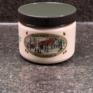 May include: A white jar with a brown lid containing pure maple cream. The jar has a label with a red maple leaf and a picture of a cabin in the woods. The label reads "Pure maple cream" and "226.8 g Half Lb."