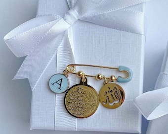 May include: Gold safety pin with charms. The charms include a round charm with the word "Allah" in Arabic script, a round charm with Arabic script, and a round charm with the letter "A" in white on a blue background.