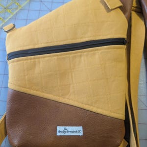 Op de afbeelding: Mosterdgele en bruine sling bag met een zwarte rits. De tas heeft een gewatteerde textuur op de gele stof en een bruin, leerachtig materiaal aan de onderkant. De tas heeft een riem en een klein label met de tekst "The Hawley Homestead LLC".