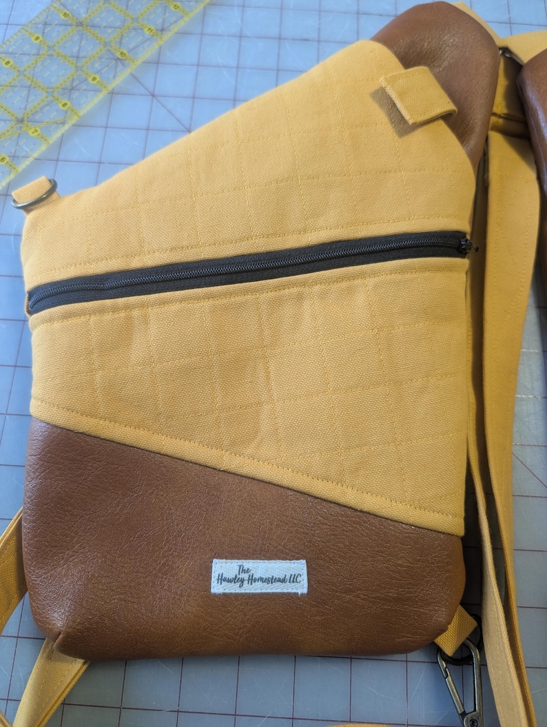 Op de afbeelding: Mosterdgele en bruine sling bag met een zwarte rits. De tas heeft een gewatteerde textuur op de gele stof en een bruin, leerachtig materiaal aan de onderkant. De tas heeft een riem en een klein label met de tekst "The Hawley Homestead LLC".