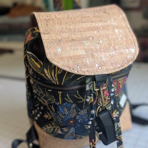 May include: A floral print backpack with a cork flap closure. The backpack features a black base with a colorful floral pattern, a cork flap with silver accents, and black straps. The bag has a zippered front pocket and adjustable straps.