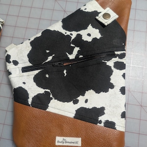 Op de afbeelding: Een crossbodytas met een koeienprint. De tas heeft een zwarte rits, bruine kunstlederen accenten en een riem. De tas heeft een label met de tekst "The Hawley Homestead LLC".