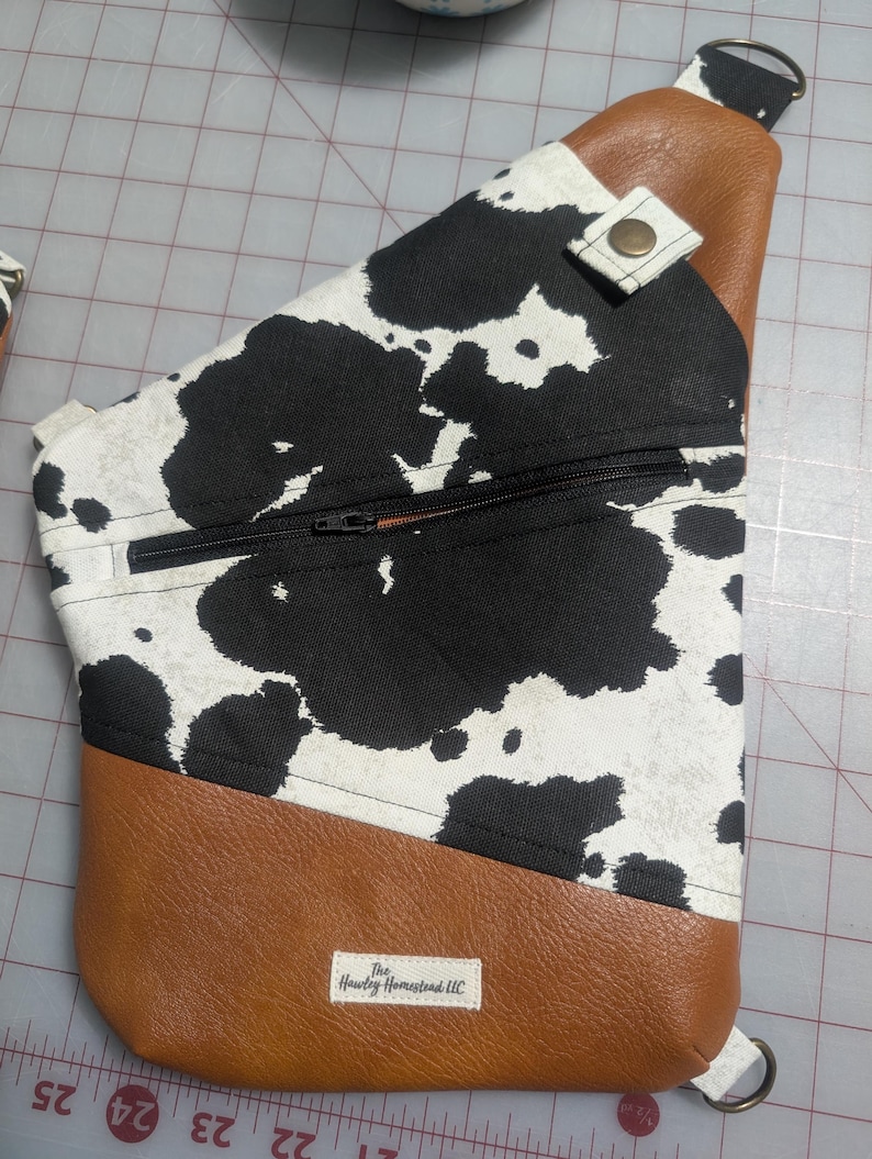 Op de afbeelding: Een crossbodytas met een koeienprint. De tas heeft een zwarte rits, bruine kunstlederen accenten en een riem. De tas heeft een label met de tekst "The Hawley Homestead LLC".