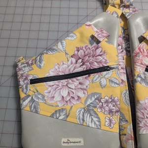 Op de afbeelding: Een gele sling bag met een bloemenpatroon en een groot roze en grijs bloemenpatroon. De tas heeft een grijs onderste gedeelte en een zwarte rits. De tas heeft verstelbare bandjes en een klein label "The Hawley Homestead LLC".