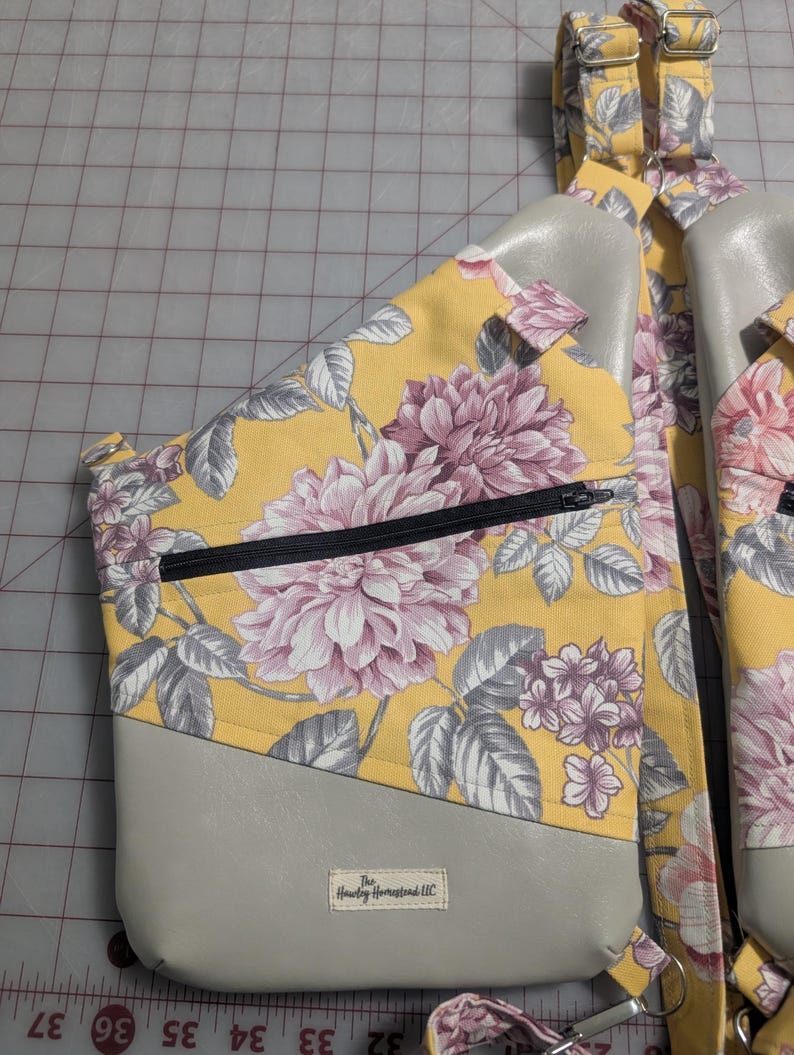 Op de afbeelding: Een gele sling bag met een bloemenpatroon en een groot roze en grijs bloemenpatroon. De tas heeft een grijs onderste gedeelte en een zwarte rits. De tas heeft verstelbare bandjes en een klein label "The Hawley Homestead LLC".