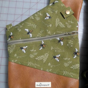 Op de afbeelding: Olijfgroene crossbodytas met een bijen- en bloemenpatroon. De tas heeft een bruine kunstleren onderkant en een vak met ritssluiting. De tas heeft een riem en een drukknoopsluiting. Het label luidt "The Hawley Homestead LLC".