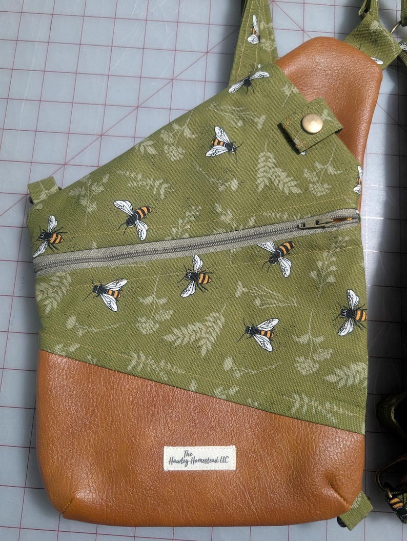 Op de afbeelding: Olijfgroene crossbodytas met een bijen- en bloemenpatroon. De tas heeft een bruine kunstleren onderkant en een vak met ritssluiting. De tas heeft een riem en een drukknoopsluiting. Het label luidt "The Hawley Homestead LLC".