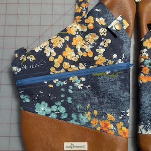 Op de afbeelding: Een marineblauwe sling bag met een bloemenpatroon in geel en wit. De tas heeft een bruine, lederlook bodem en een blauwe rits. De riem is ook voorzien van een geel en wit bloemendesign. De tas heeft een klein label met "The Hawley Homestead LLC."