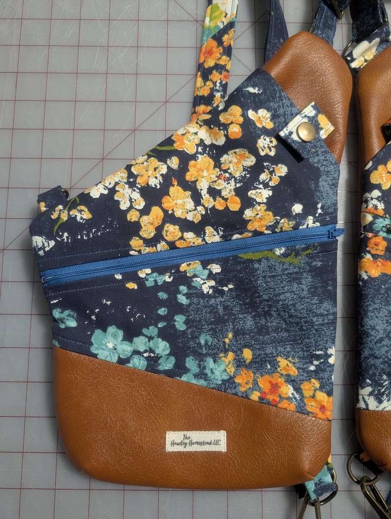 Op de afbeelding: Een marineblauwe sling bag met een bloemenpatroon in geel en wit. De tas heeft een bruine, lederlook bodem en een blauwe rits. De riem is ook voorzien van een geel en wit bloemendesign. De tas heeft een klein label met "The Hawley Homestead LLC."