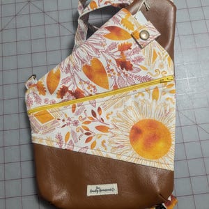 Op de afbeelding: Bruin-witte sling bag met een zonnebloemontwerp en een gele rits. De tas heeft een bruine imitatielederen bodem en riem, en een bloemenpatroon met oranje en gele accenten. De tas heeft een klein label met "The Howdy Homestead Co."