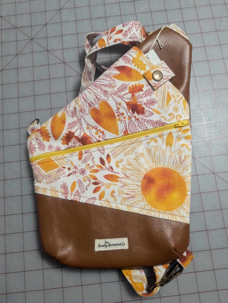 Op de afbeelding: Bruin-witte sling bag met een zonnebloemontwerp en een gele rits. De tas heeft een bruine imitatielederen bodem en riem, en een bloemenpatroon met oranje en gele accenten. De tas heeft een klein label met "The Howdy Homestead Co."