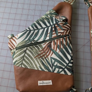 Op de afbeelding: Een bruine en groene sling bag met een tropisch bladerpatroon. De tas heeft een bruine leren basis en riem, een groene rits en een wit label met tekst. De tas is ontworpen om over het lichaam te worden gedragen.