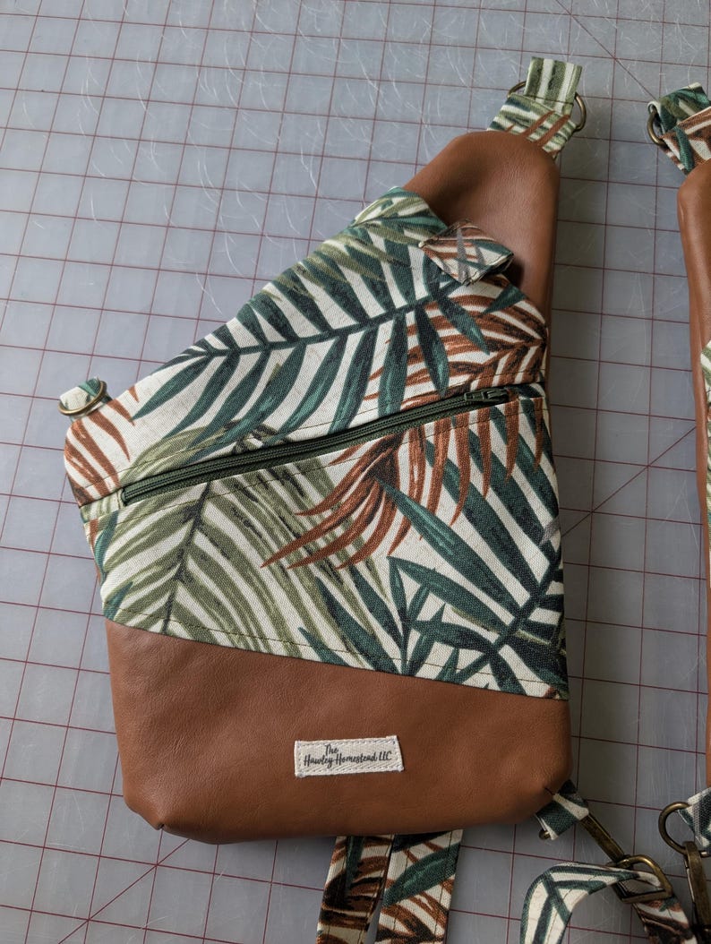 Op de afbeelding: Een bruine en groene sling bag met een tropisch bladerpatroon. De tas heeft een bruine leren basis en riem, een groene rits en een wit label met tekst. De tas is ontworpen om over het lichaam te worden gedragen.