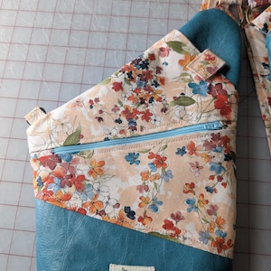 Op de afbeelding: Een teal en bloemen sling bag met een lichtblauwe rits. De tas heeft een bloemenpatroon met oranje, rode en blauwe bloemen op een perzik achtergrond. De onderste sectie is teal, en de riem heeft een bijpassend bloemenontwerp.