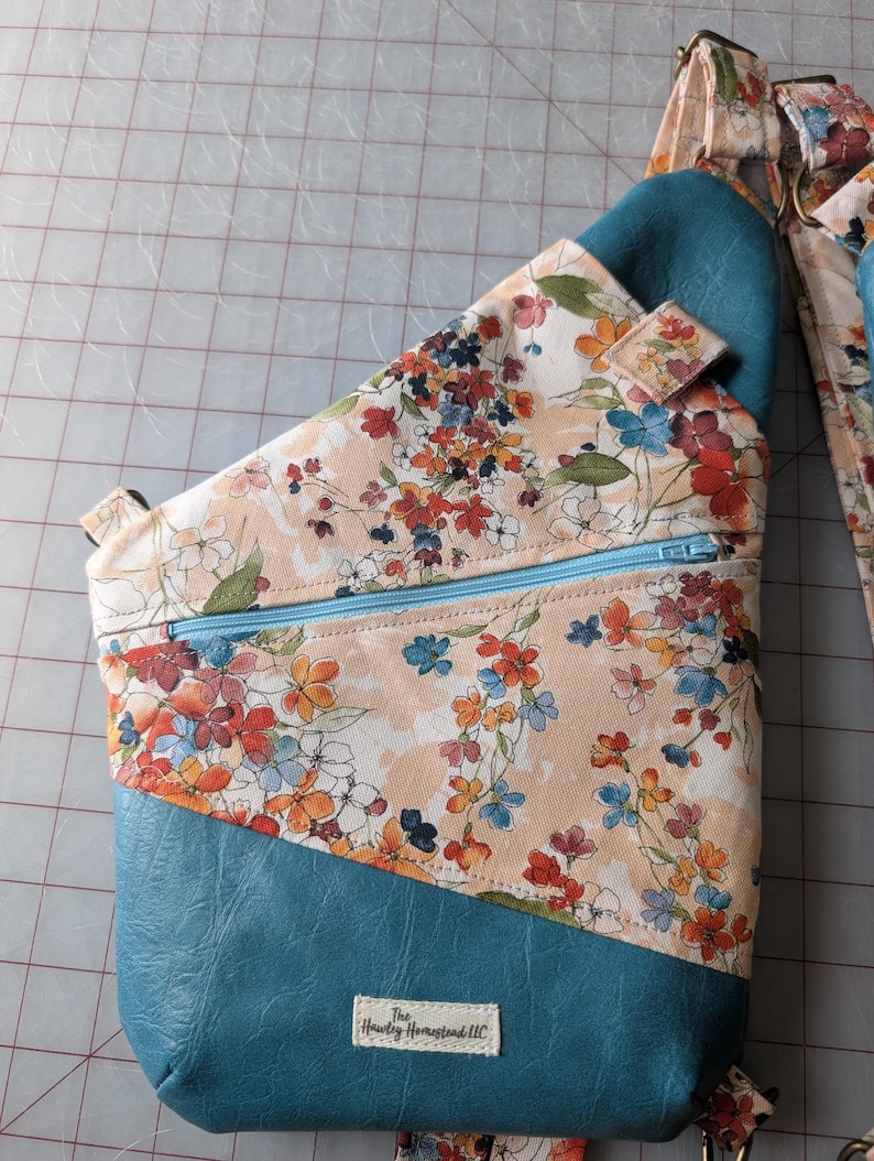 Op de afbeelding: Een teal en bloemen sling bag met een lichtblauwe rits. De tas heeft een bloemenpatroon met oranje, rode en blauwe bloemen op een perzik achtergrond. De onderste sectie is teal, en de riem heeft een bijpassend bloemenontwerp.