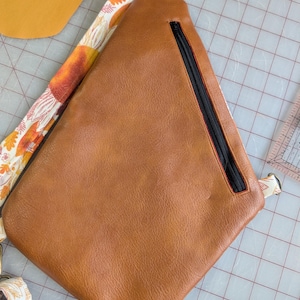 Op de afbeelding: Een bruine leren sling bag met een zwarte rits en een band met oranje en witte bloemmotieven. De tas ligt op een rasteroppervlak, met een geel stuk stof zichtbaar.