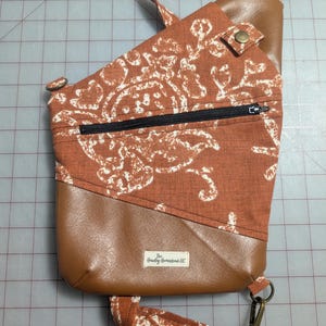 Op de afbeelding: Een crossbodytas met een bovenkant van roestkleurige stof met een wit bloemmotief. Het onderste gedeelte en de accenten zijn van bruin imitatieleer. Het heeft een zwarte ritssluiting, een drukknoopsluiting en een riem met een clip.