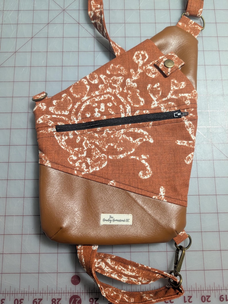 Op de afbeelding: Een crossbodytas met een bovenkant van roestkleurige stof met een wit bloemmotief. Het onderste gedeelte en de accenten zijn van bruin imitatieleer. Het heeft een zwarte ritssluiting, een drukknoopsluiting en een riem met een clip.