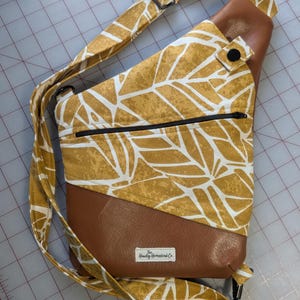 Op de afbeelding: Een mosterdgele sling bag met een wit bladerenpatroon en bruine, lederachtige accenten. De tas heeft een zwarte ritssluiting, een verstelbare riem en een label met "Hawley Homestead Co."